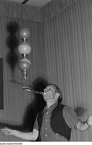 Artist_Bälle_auf_einem_Messer_balancierend man balancing balls and glasses on a rod from his mouth