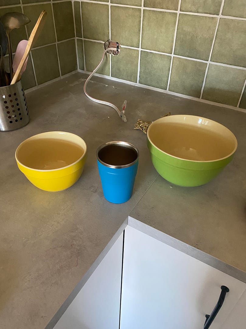 image shows three objects on a concrete coloured bench top with green tiles, moon white grout and electrical works behind. The three bowls are a yellow small bowl, a medium blue keep cup and a larger green mixing bowl (ceramic)